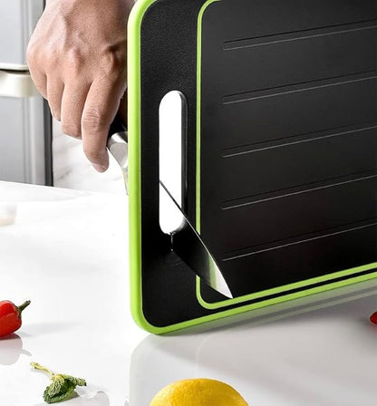 Person using a green-edged double-sided chopping board for cutting, with knife slot for safety, surrounded by fresh vegetables.